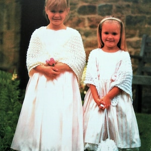May include: Two young girls wearing white dresses and crocheted shawls. The girl on the left is holding a bouquet of pink flowers. The girl on the right is holding a white crocheted bag.