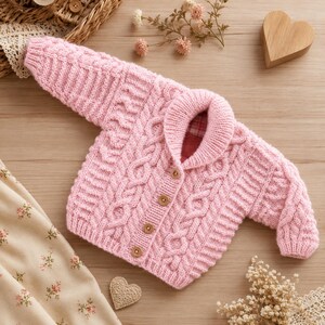 May include: A light pink baby cardigan with a cable knit design. The sweater has a shawl collar, wooden buttons, and ribbed cuffs. The interior collar lining has a plaid pattern. The garment is displayed on a wooden surface.