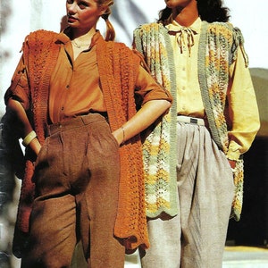 May include: Two women wearing crocheted vests. The woman on the left is wearing a brown vest with a brown shirt and brown trousers. The woman on the right is wearing a green, white and orange vest with a yellow shirt and beige trousers.