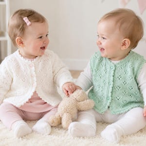 May include: Two infants sitting together, one wearing a white long-sleeved knit cardigan with a delicate eyelet pattern and small buttons, the other wearing a light green sleeveless knit vest with a matching eyelet pattern and buttons.
