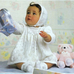 Puede incluir: Un bebé con un vestido, gorro y calcetines de punto blanco, sosteniendo un regalo envuelto. El vestido tiene un patrón decorativo y un lazo en la cintura. Un osito de peluche rosa está cerca. El fondo presenta un papel pintado con diseños de animales pastel.