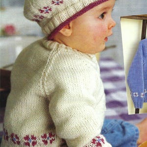 May include: A white knitted baby sweater with a pink and blue floral pattern on the cuffs and hem. The sweater is worn by a baby wearing a white knitted hat with a pink trim and a white pom-pom.