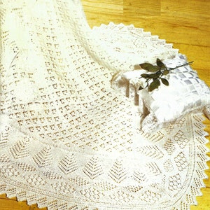 Peut inclure: Une couverture de bébé en crochet blanc avec un motif de dentelle délicat. La couverture est posée sur un sol en bois avec un oreiller blanc et une brindille de verdure.