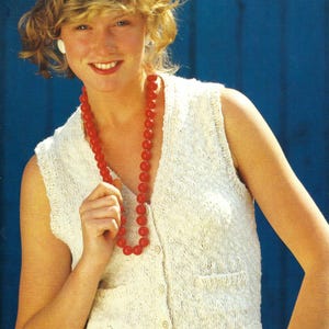 Peut inclure: Un gilet sans manches en tricot blanc avec une fermeture boutonnée et deux poches. Le gilet est porté sur une chemise blanche et associé à un béret rouge et un collier de perles rouges.