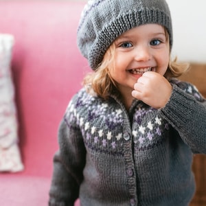 May include: A grey knitted cardigan and matching hat with a pom-pom. The sweater and hat have a white and purple floral pattern. The child is smiling and wearing a grey knitted beret.