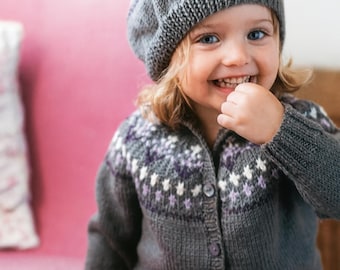May include: A gray knit cardigan and matching hat with a pom-pom. The sweater and hat have a white and purple floral pattern. The child is smiling and wearing a gray knit beret.