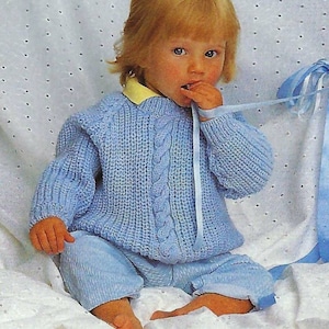 May include: A baby wearing a light blue knitted sweater with a cable knit design and matching pants. The sweater has a round neckline and long sleeves. The baby is sitting on a white blanket and holding a blue ribbon.