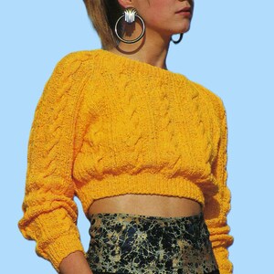 May include: A cropped, bright yellow cable-knit sweater with long sleeves. The model is wearing large hoop earrings and a patterned belt. The sweater is paired with black trousers. The background is a light blue.