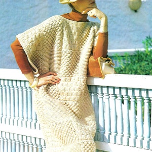 May include: A white crocheted poncho with a textured pattern. The poncho is being worn by a person wearing a brown turtleneck sweater and a brown hat. The person is standing in front of a white railing.