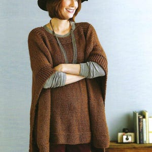 Pode incluir: Um poncho de malha marrom com decote largo e mangas compridas. O poncho está sendo usado por uma pessoa com cabelo castanho e um chapéu preto. A pessoa está usando uma camisa cinza de manga comprida por baixo do poncho.