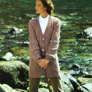 May include: A woman wearing a long, brown cable knit cardigan with a white button-down shirt underneath. She is standing in a natural setting with a river and rocks in the background.