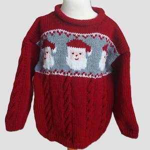 May include: A red knitted Christmas jumper with a grey panel featuring Santa Claus faces. The jumper has a cable knit design on the lower section and sleeves. The collar is a darker shade of red.