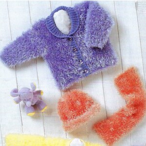Puede incluir: Un cárdigan de punto morado y peludo con cierre de botones, un gorro y una bufanda a juego en naranja, y un elefante de peluche morado.