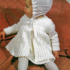 May include: A white knitted baby set consisting of a long-sleeved cardigan with a ribbed skirt and a matching bonnet. The cardigan features a front ribbon tie closure. The set is paired with white patterned tights featuring a diamond-shaped texture.