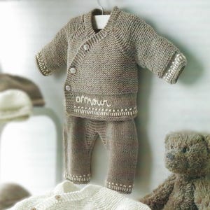 May include: A knitted baby outfit in a light brown colour with a white embroidered word "amour" on the front. The outfit includes a cardigan with a button closure and matching trousers.