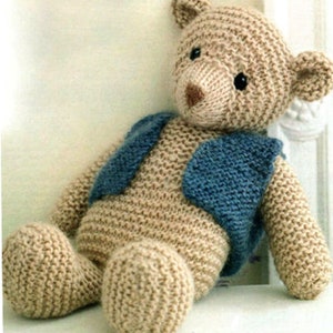 May include: A knitted teddy bear with a blue vest. The bear is sitting on a white surface.