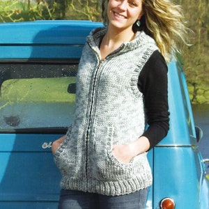 May include: A woman wearing a gray hooded knitted vest with a zipper and pockets, standing next to a blue car.