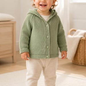 Op de afbeelding: Een saliegroen gebreid vest met capuchon, voorzien van een knoopsluiting aan de voorzijde met vier bijpassende knopen, lange mouwen en een geribbelde zoom. Het vest wordt gedragen over een effen wit shirt met een lichtbeige broek en witte sneakers.
