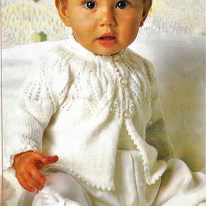 May include: A white knitted baby cardigan with a button closure. The cardigan has a delicate lace pattern and is worn by a baby sitting on a white blanket.