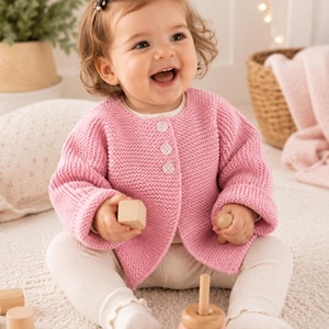 Può includere: Un bambino sorridente con un cardigan rosa chiaro lavorato a maglia con tre bottoni bianchi, abbinato a leggings bianchi a coste. Il bambino tiene in mano un blocchetto di legno e siede su un soffice tappeto bianco, con giocattoli di legno nelle vicinanze.