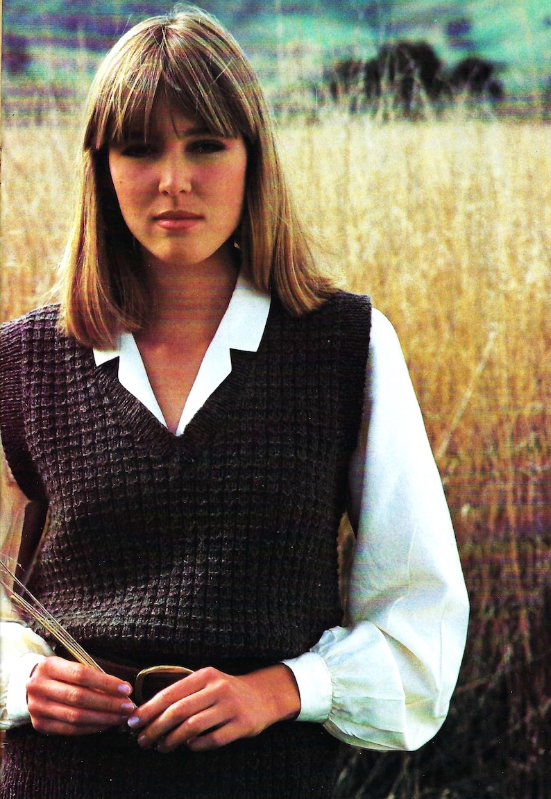 Op de afbeelding: Een vrouw draagt een donkerbruin gebreid vest met daaronder een wit overhemd met kraag. Het vest heeft een textuurpatroon. De achtergrond is een veld met hoog, droog gras.