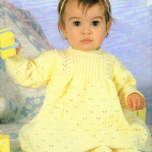May include: A baby girl wearing a yellow knitted dress with a delicate lace pattern. She is holding a small gift box and wearing a yellow headband.
