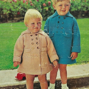 May include: Two children wearing knitted coats. The girl is wearing a beige coat with brown buttons and the boy is wearing a blue coat with brown buttons. Both children are wearing white socks and white shoes.