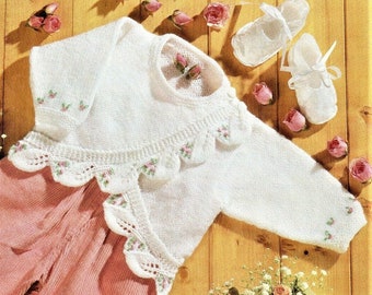 May include: White knitted baby sweater with a scalloped edge and floral embroidery. The sweater is laid out on a wooden surface with pink roses and white baby shoes.