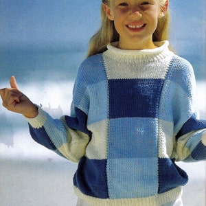 May include: A young person wearing a white, blue, and light blue checkered sweater. The sweater has a high neckline and long sleeves. The person is smiling and giving a thumbs up.