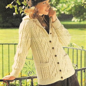 May include: A cream-colored knitted cardigan with a cable knit pattern, a hood, and a button closure. The cardigan has two patch pockets and is worn by a person with brown hair and a brown beret.