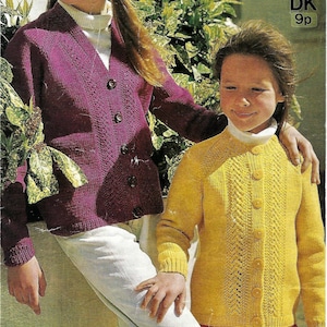 May include: Two girls wearing knitted cardigans. The girl on the left is wearing a purple cardigan with a button-up front. The girl on the right is wearing a yellow cardigan with a button-up front. Both cardigans have a cable knit pattern.