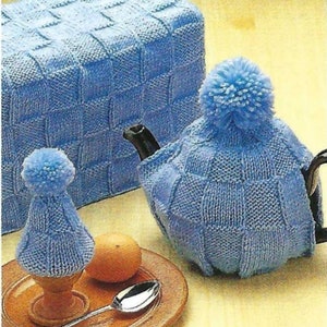 Puede incluir: Un conjunto de tres cubiertas de punto azul para una tetera, un huevo y una caja de pan. Las cubiertas están hechas con un punto de punto simple y tienen un pompón en la parte superior.