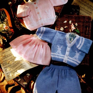 Może przedstawiać: Różowy dzianinowy kardigan z białymi lamówkami i guzikami, różowa dzianinowa spódnica, niebieski dzianinowy kardigan z białymi lamówkami i guzikami oraz niebieskie dzianinowe spodnie. Ubrania leżą na powierzchni z nutami i wiklinowym koszem.