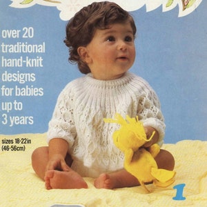 Puede incluir: Un jersey de punto blanco para bebé con un delicado patrón de encaje. El jersey lo lleva un bebé sentado en una manta amarilla, sosteniendo un juguete de peluche amarillo. El texto de la página dice "más de 20 diseños tradicionales tejidos a mano para bebés de hasta 3 años tallas 18-22 pulgadas (46-56 cm)"
