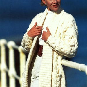 May include: A white knitted cardigan with a cable knit pattern and a long, open front. The cardigan is being worn by a person with short blonde hair and is standing in front of a blue background.