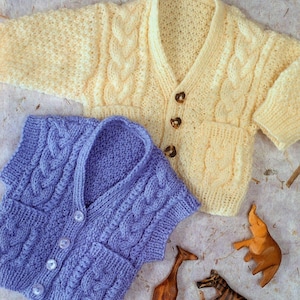 Könnte beinhalten: Zwei Strickjacken, eine gelbe und eine lila, mit Zopfmuster und Knöpfen. Die Jacken liegen auf einer strukturierten Oberfläche mit Holztierfiguren.