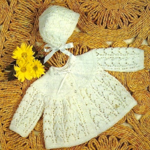 Puede incluir: Un gorro y un cárdigan de bebé de ganchillo blanco con un delicado patrón de encaje. El conjunto se coloca sobre un fondo de paja tejida.