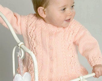 May include: A baby girl wearing a pink knitted cardigan with a button closure and a delicate lace pattern. The cardigan has long sleeves and a slightly flared hem. The baby is holding onto a white metal crib.