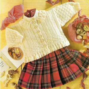 May include: A cream-coloured knitted cardigan with heart-shaped buttons, paired with a red and black plaid pleated skirt. The cardigan has textured sleeves and a ruffled hem. A red and white striped scarf and a basket with shells complete the ensemble.