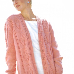 May include: A light pink cardigan sweater with a diamond and leaf pattern. The sweater has long sleeves and a button-up front. The cardigan is made of a soft, knitted material. The model is wearing a white top underneath.