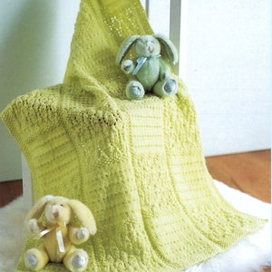 May include: A light green knitted baby blanket with a delicate stitch pattern. Two plush bunny toys are sitting on the blanket, one green and one cream.