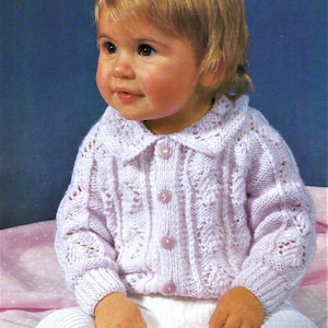 May include: A pink knitted baby cardigan with a white collar and button closure. The cardigan has a delicate lace pattern.