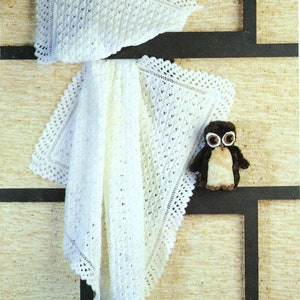 Puede incluir: Una manta de bebé de ganchillo blanco con un delicado borde de encaje. La manta está doblada por la mitad y drapeada sobre una superficie marrón. Un pequeño búho de peluche está sentado en la superficie.