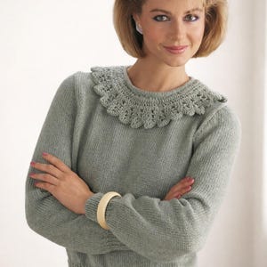 May include: A light green sweater with a crocheted collar. The long-sleeved sweater has a ribbed hem and cuffs. A cream-colored bracelet is visible on the left wrist. The woman is wearing earrings and has short, styled hair.