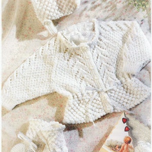 Puede incluir: Un conjunto de ropa de bebé de punto blanco, que incluye un cárdigan, un gorro y botines. El cárdigan tiene un delicado patrón de encaje y un cierre de lazo. El gorro y los botines también están tejidos con un punto texturizado.