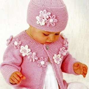 Peut inclure: Un cardigan rose en tricot pour bébé avec des fleurs blanches en embellissement et un bonnet rose en tricot assorti avec des fleurs blanches en embellissement.