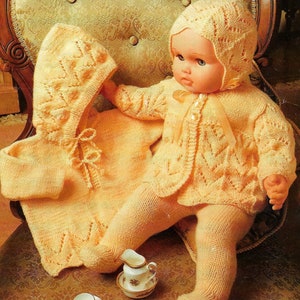 May include: A knitted baby doll outfit in a pale yellow color. The outfit includes a hooded jacket, a cardigan, and leggings. The doll is sitting on a chair with a floral pattern.