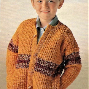 Op de afbeelding: Een jonge jongen draagt een gele gebreide cardigan met bruine strepen. De cardigan heeft een knoopsluiting aan de voorkant en zakken aan de zijkanten.