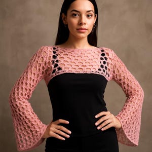 Op de afbeelding: Een lichtroze, open geweven gehaakte bolero met lange mouwen. De bolero wordt over een zwarte top gedragen. De mouwen vallen wijd uit bij de polsen. Het kledingstuk heeft een delicate, luchtige uitstraling, geschikt om in laagjes te dragen.