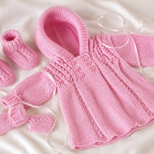 May include: A hand-knitted baby set in light pink, consisting of a long-sleeved hooded cardigan with a button-front closure, a pair of matching booties with ribbed cuffs, and two small mittens, all featuring a consistent cable knit pattern.
